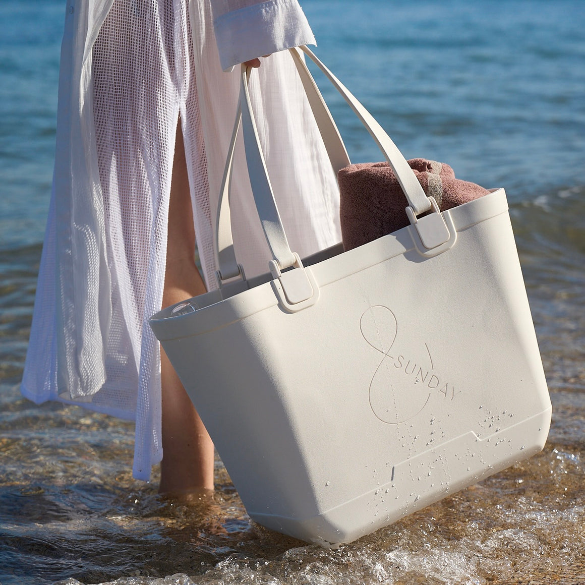 Person holding a beige tote bag with '&SUNDAY' branding at the beach.