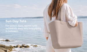 Person holding a beige tote bag by the ocean with text about the 'Sun Day Tote'.