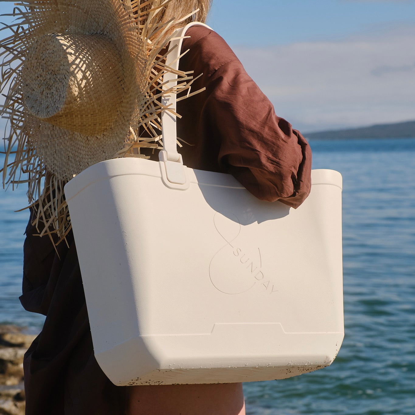 Person holding a white tote bag with a straw hat against a scenic background